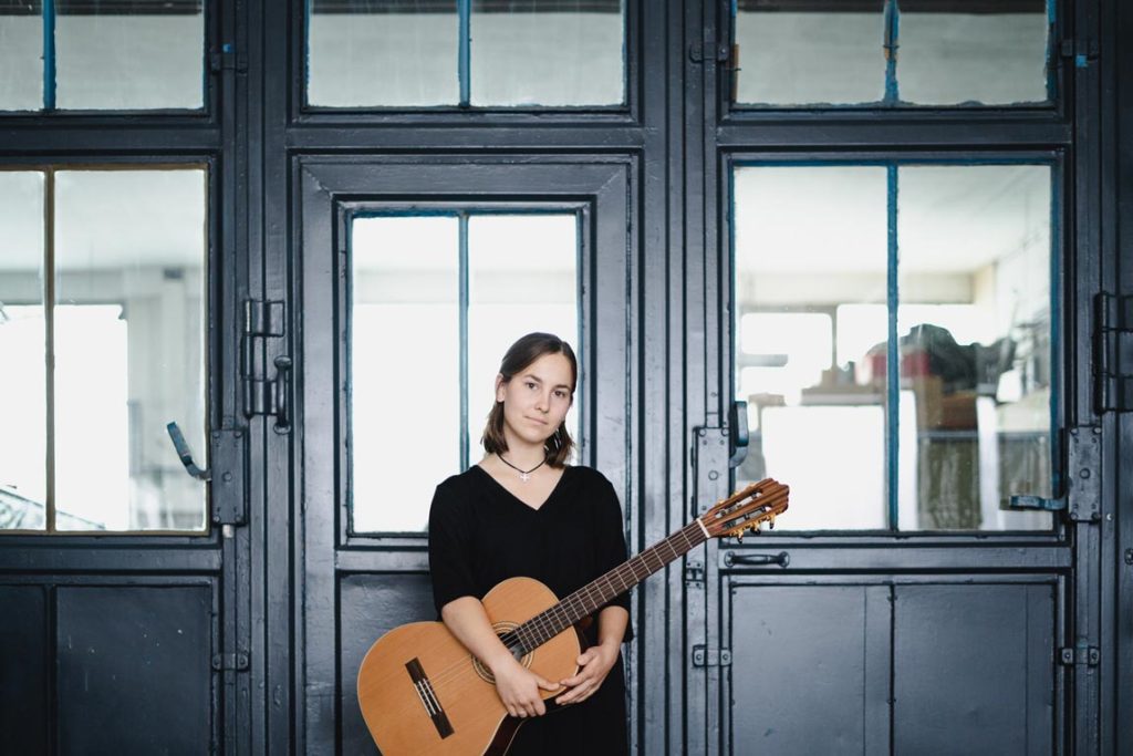 Musikerportrait Tina Wirsching steht mit Gitarre im Arm vor grauer Werkstatttür