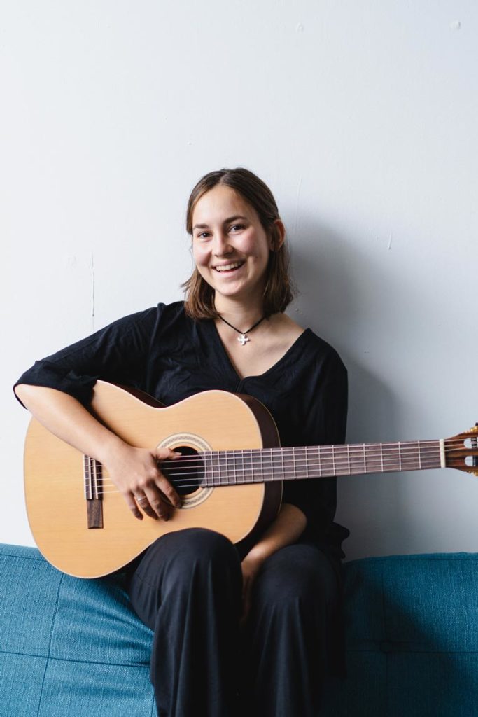Musikerportrait Tina Wirsching sitzt mit Gitarre lachen dauf der Lehne eines Sofas
